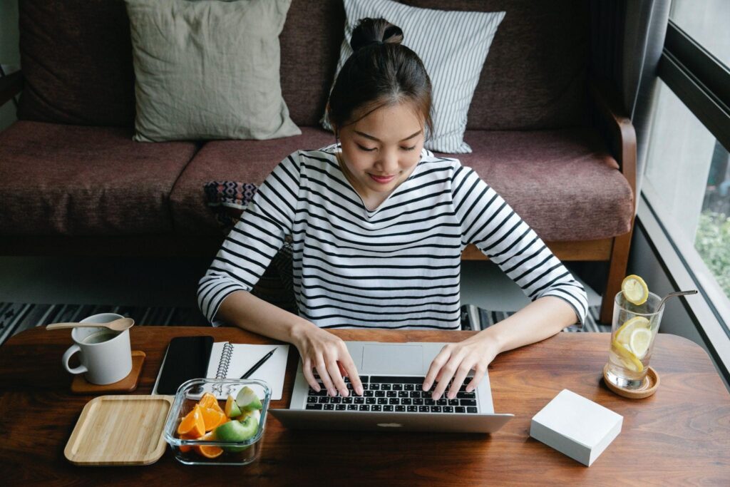 Asian freelancer typing on laptop at table with lemonade indoors